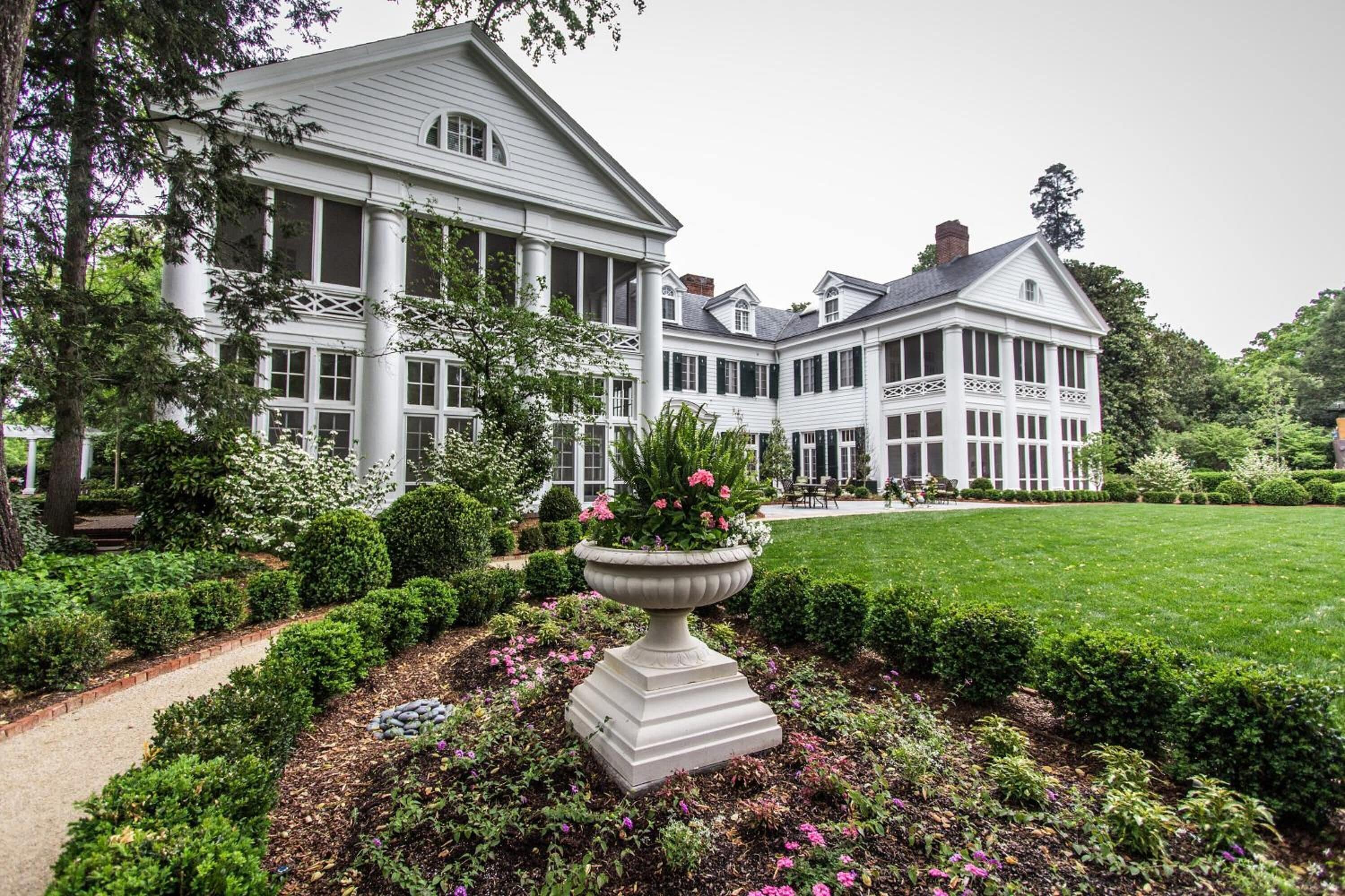 A large, white colonial-style building with multiple windows and a well-maintained garden with a central flower bed.