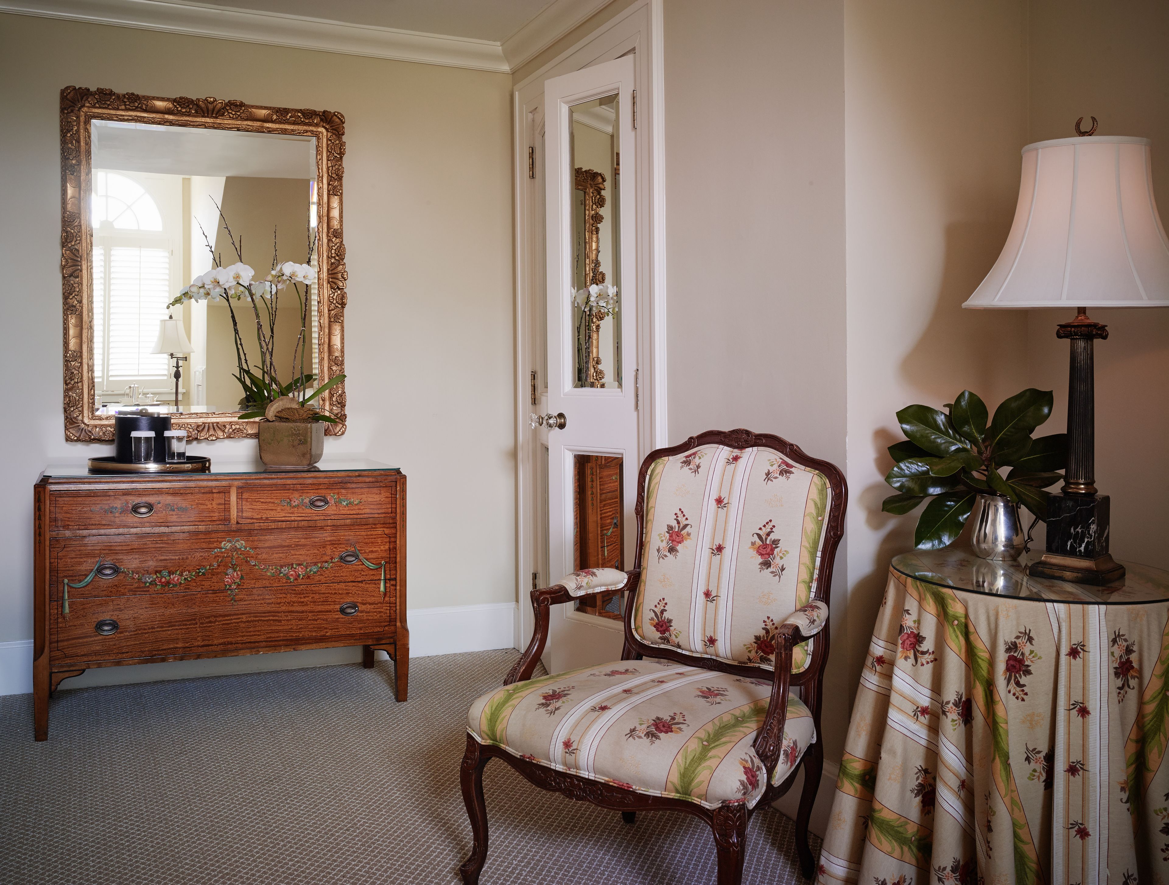 A room with a wooden dresser, a floral armchair, a mirror with an ornate frame, and a lamp on a side table.