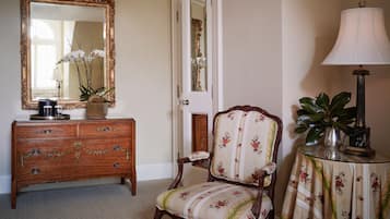 A room with a wooden dresser, a floral armchair, a mirror with an ornate frame, and a lamp on a side table.