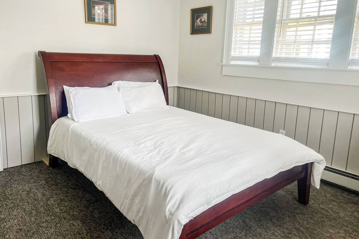 A neatly made bed with white linens in a room with a wooden headboard, white walls, and a window with blinds.