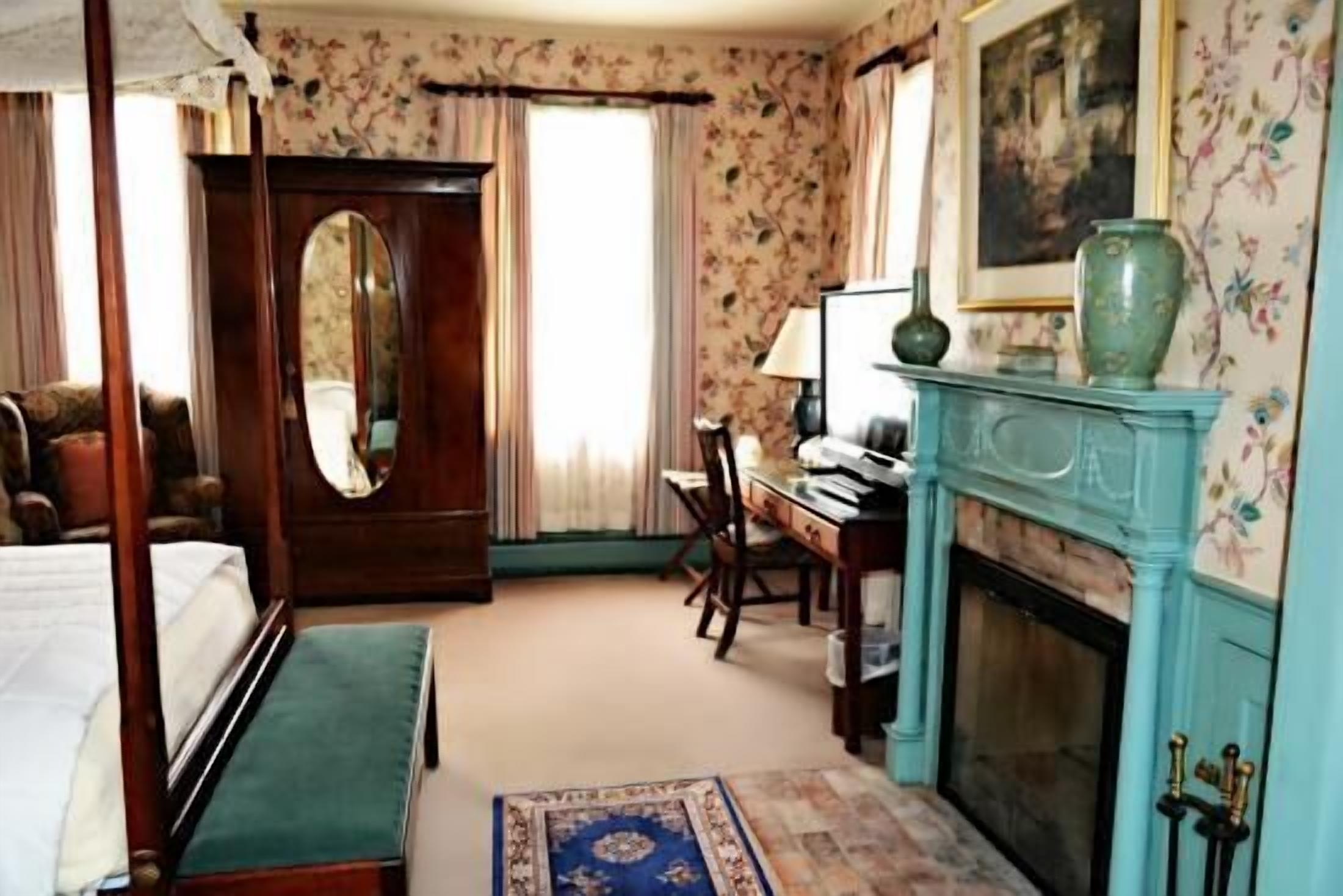 A bedroom with a four-poster bed, a desk, a chair, a fireplace, and a large mirror.