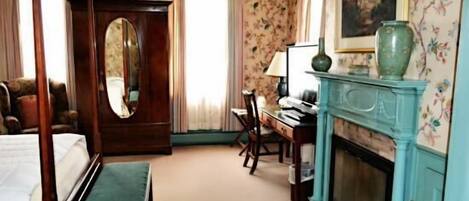 A bedroom with a four-poster bed, a desk, a chair, a fireplace, and a large mirror.