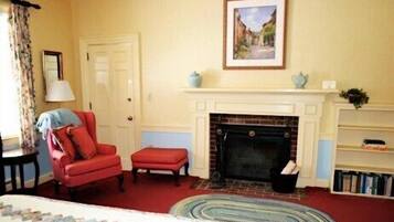 Una habitación con chimenea, un sillón rojo, un pequeño otoman, una estantería y un cuadro enmarcado en la pared.