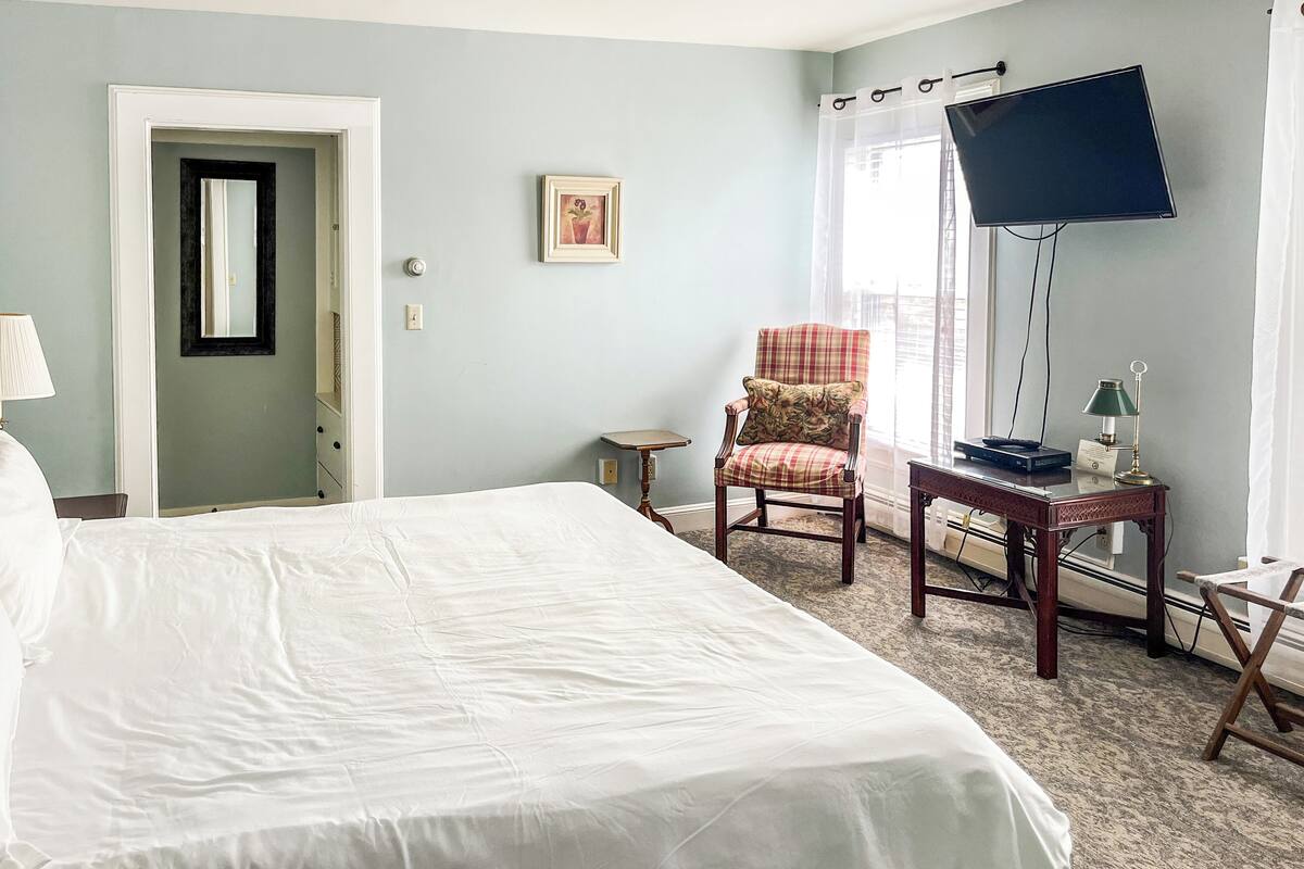 A hotel room with a large bed, a chair, a desk with a lamp, a television, and a window with curtains.