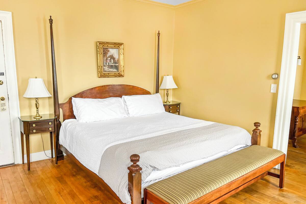 A neatly made bed with white linens, a wooden headboard, and a bench at the foot of the bed. There are two bedside tables with lamps, and a framed picture on the wall above the bed.