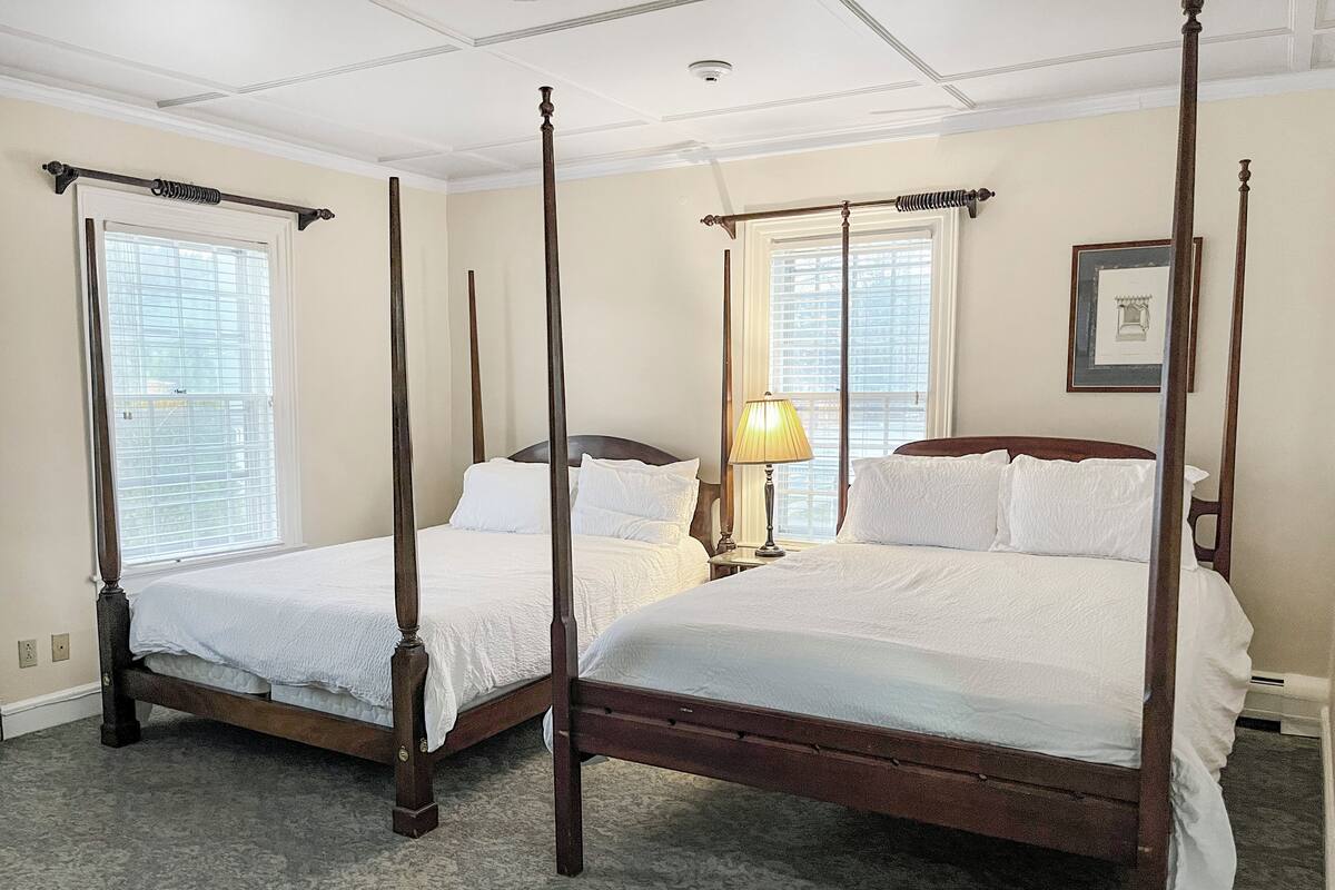 A four-poster bed with white linens in a room with two windows and a ceiling fan.
