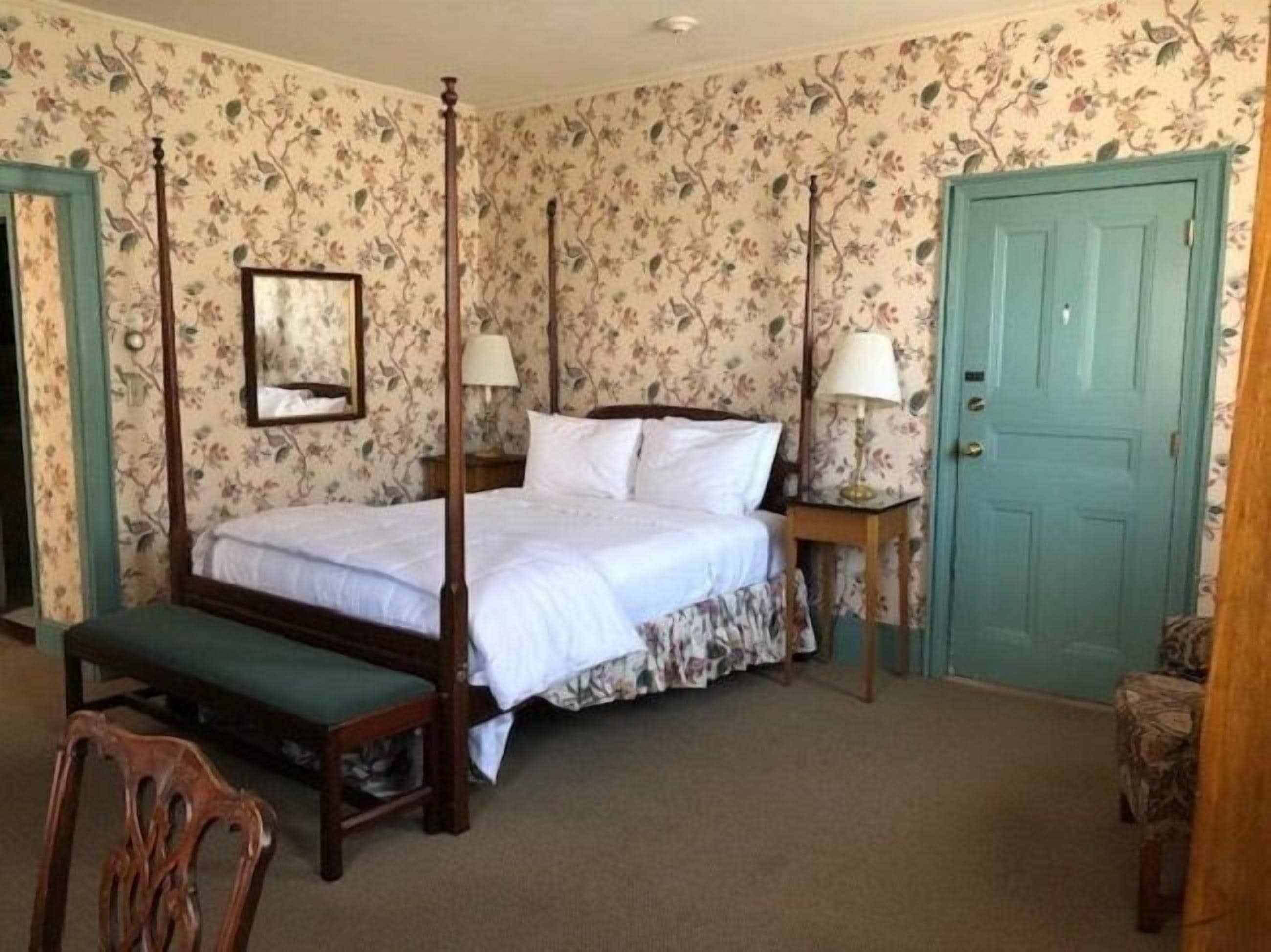 A four-poster bed with white linens, two bedside tables with lamps, a wooden bench, and a green door.