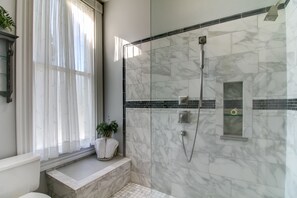 A bathroom with a walk-in shower, marble tiles, a window with sheer curtains, and a small plant on the windowsill.
