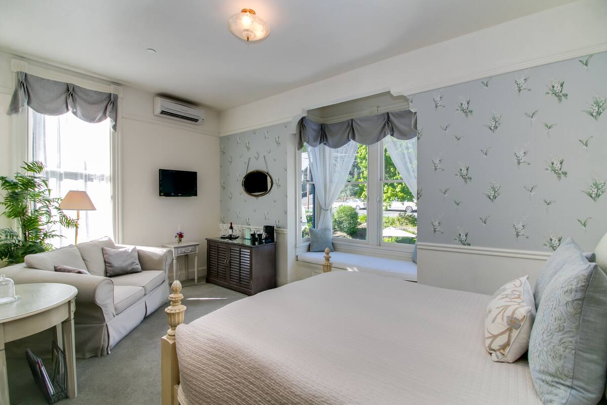 Un dormitorio con una cama grande, un sofá, una mesita y un aparador. Hay una ventana con cortinas y vistas al verdor exterior.