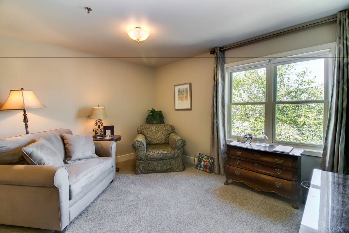Una sala de estar acogedora con un sofá color beige, un sillón estampado, una mesita con una lámpara y una ventana con cortinas.