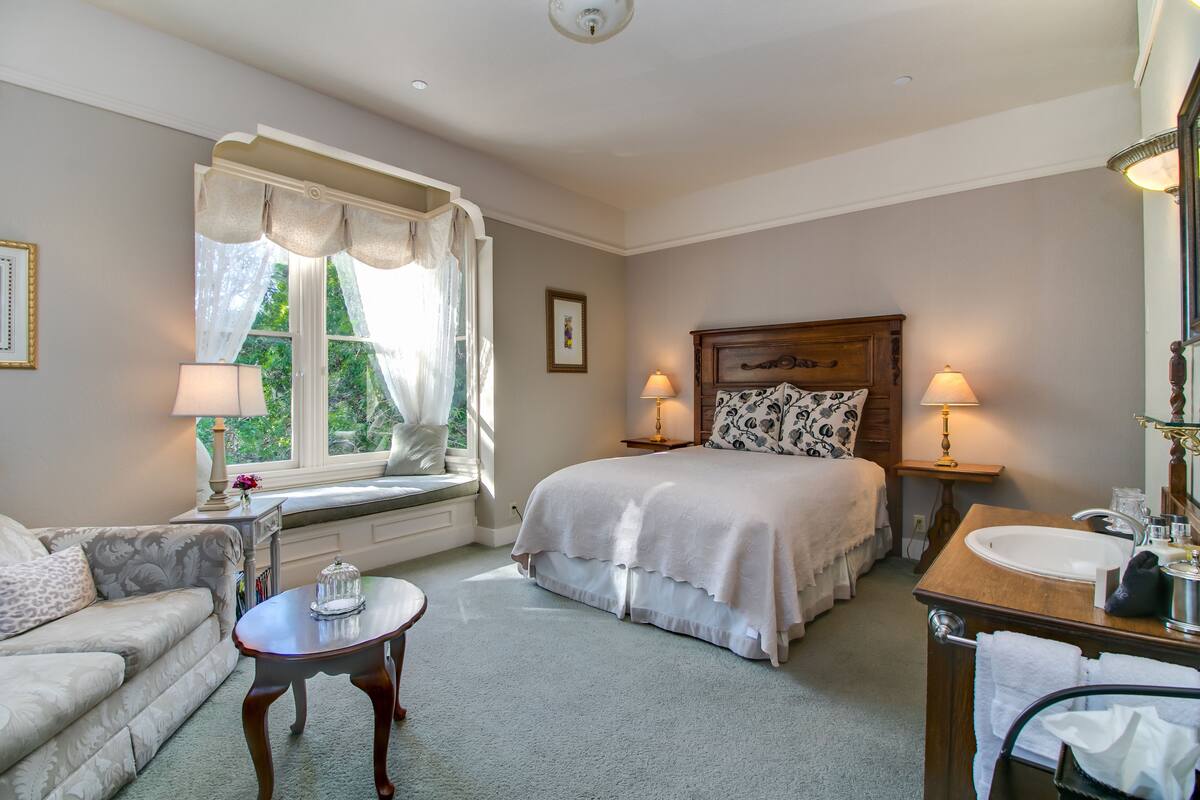 Un dormitorio con cama, lámparas de noche, ventana con cortinas, un sofá, una mesita y un baño con lavabo y espejo.