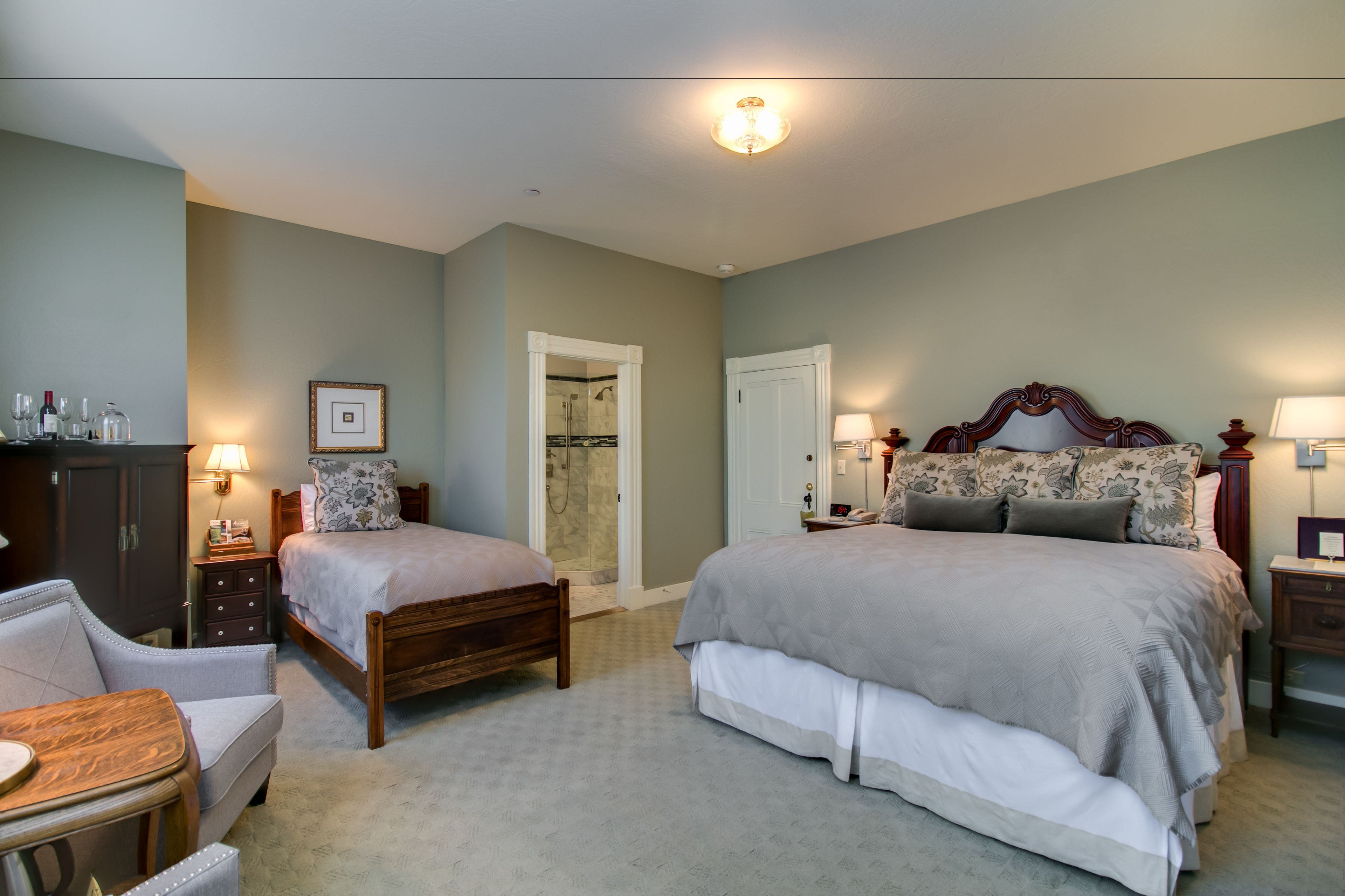 Quarto com duas camas de solteiro, uma cadeira de madeira, mesa de cabeceira com abajur e guarda-roupa.
