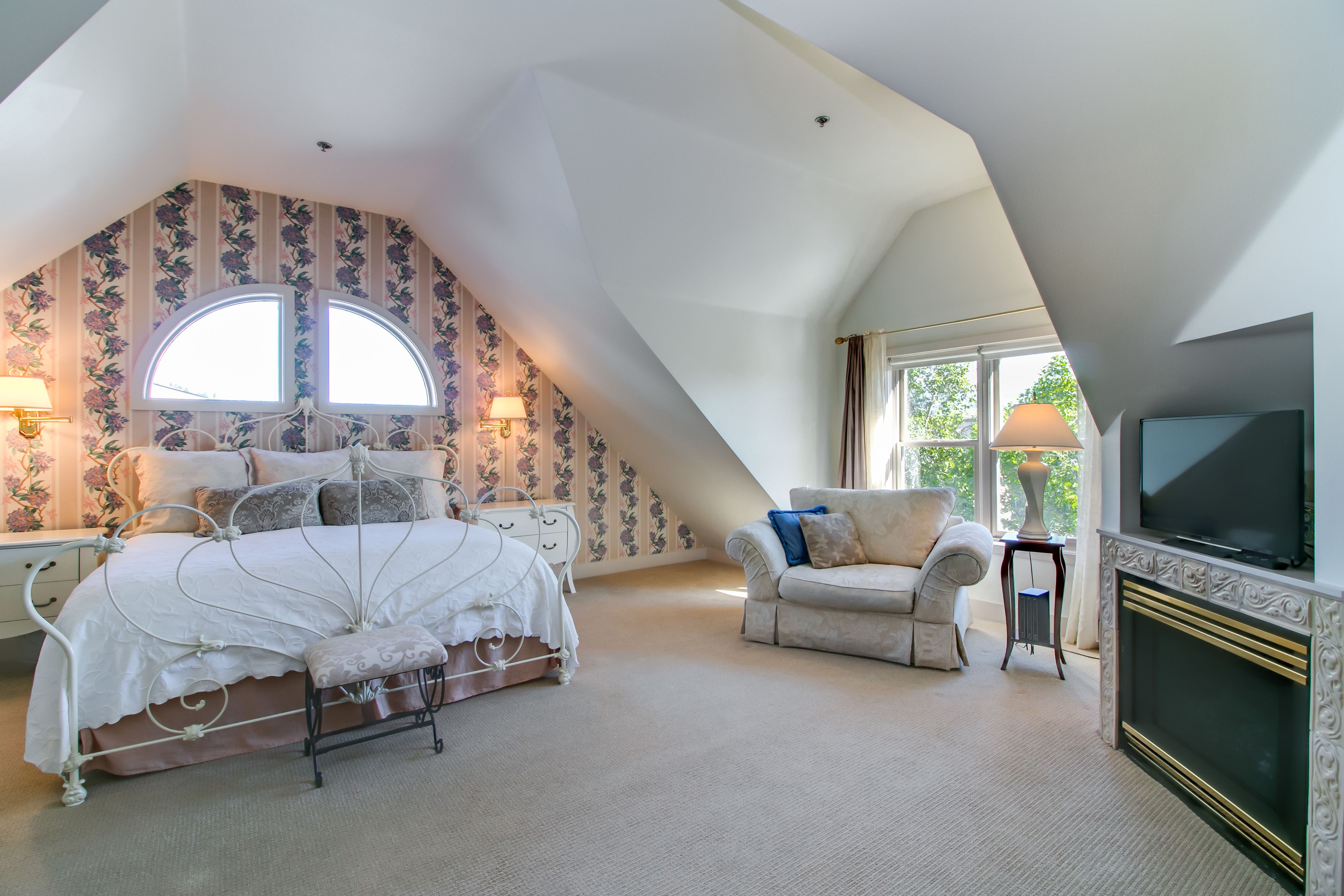 A bedroom with a sloped ceiling, a bed with a canopy, a television, a fireplace, and a window with curtains.