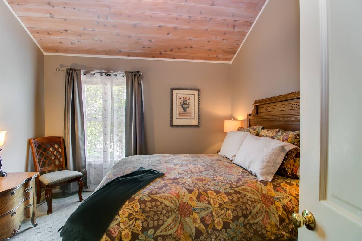 Un dormitorio con techo de madera, una cama con ropa de cama floral, una silla, un escritorio y un cuadro en la pared.