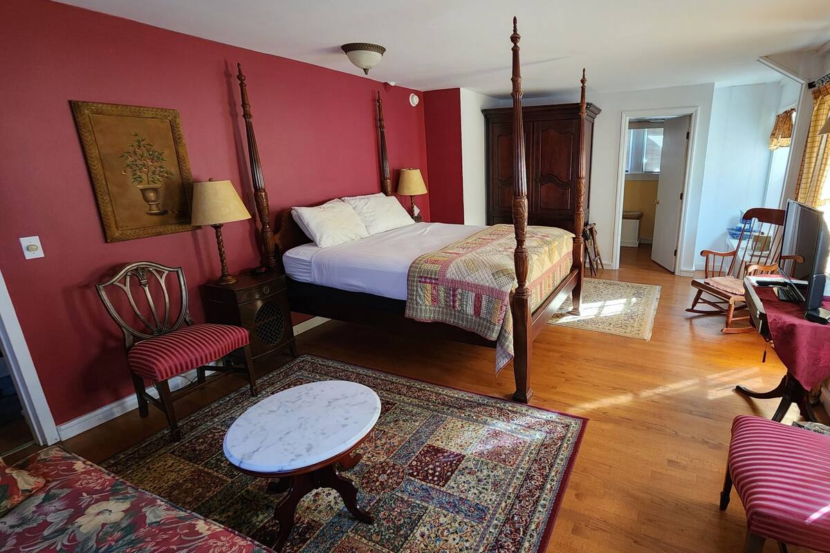Un lit à baldaquin avec une courtepointe, un fauteuil rouge rembourré, une table ronde et un tapis à motifs dans une pièce au plancher de bois et aux murs rouges.
