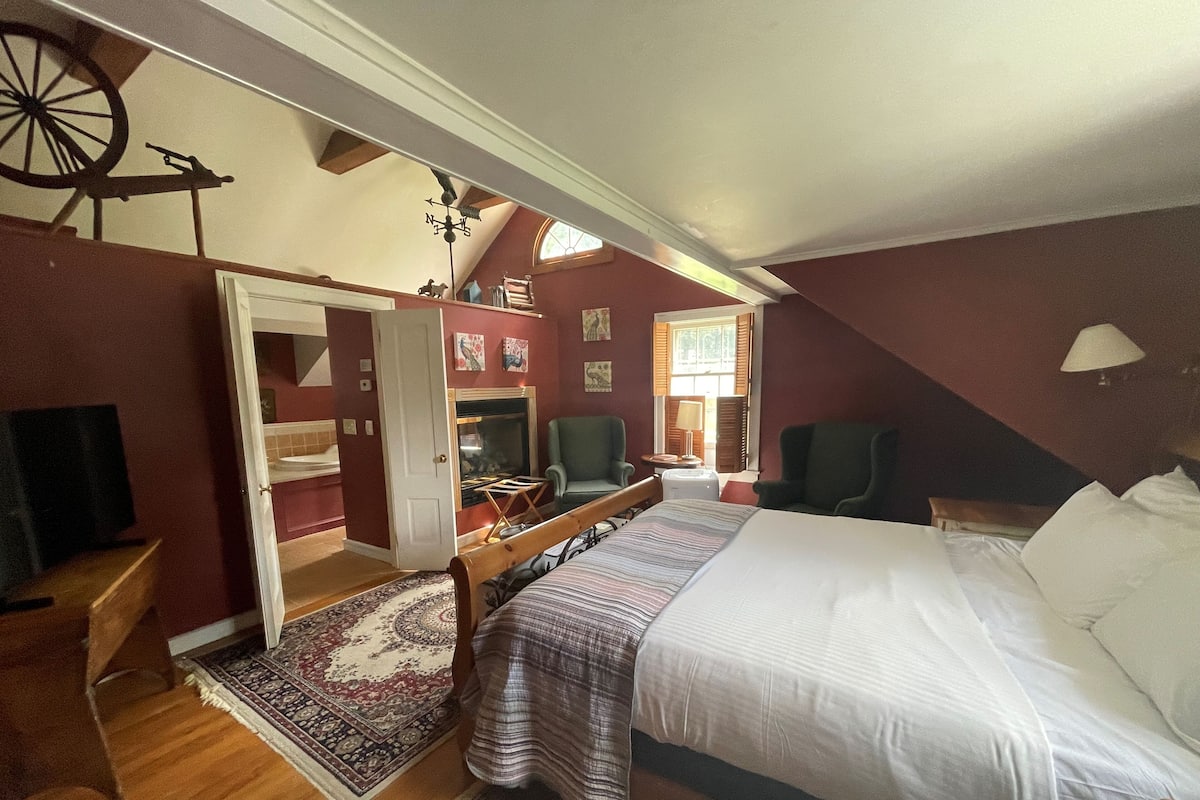 Une chambre à coucher avec un grand lit, deux fauteuils, une petite table, une cheminée et une fenêtre avec des volets.
