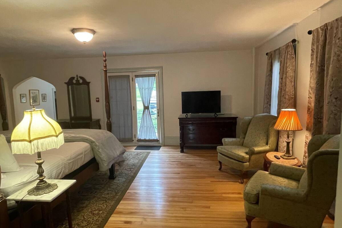 Une chambre d’hôtel avec un lit, deux fauteuils, une télévision, une commode et une fenêtre avec des rideaux.