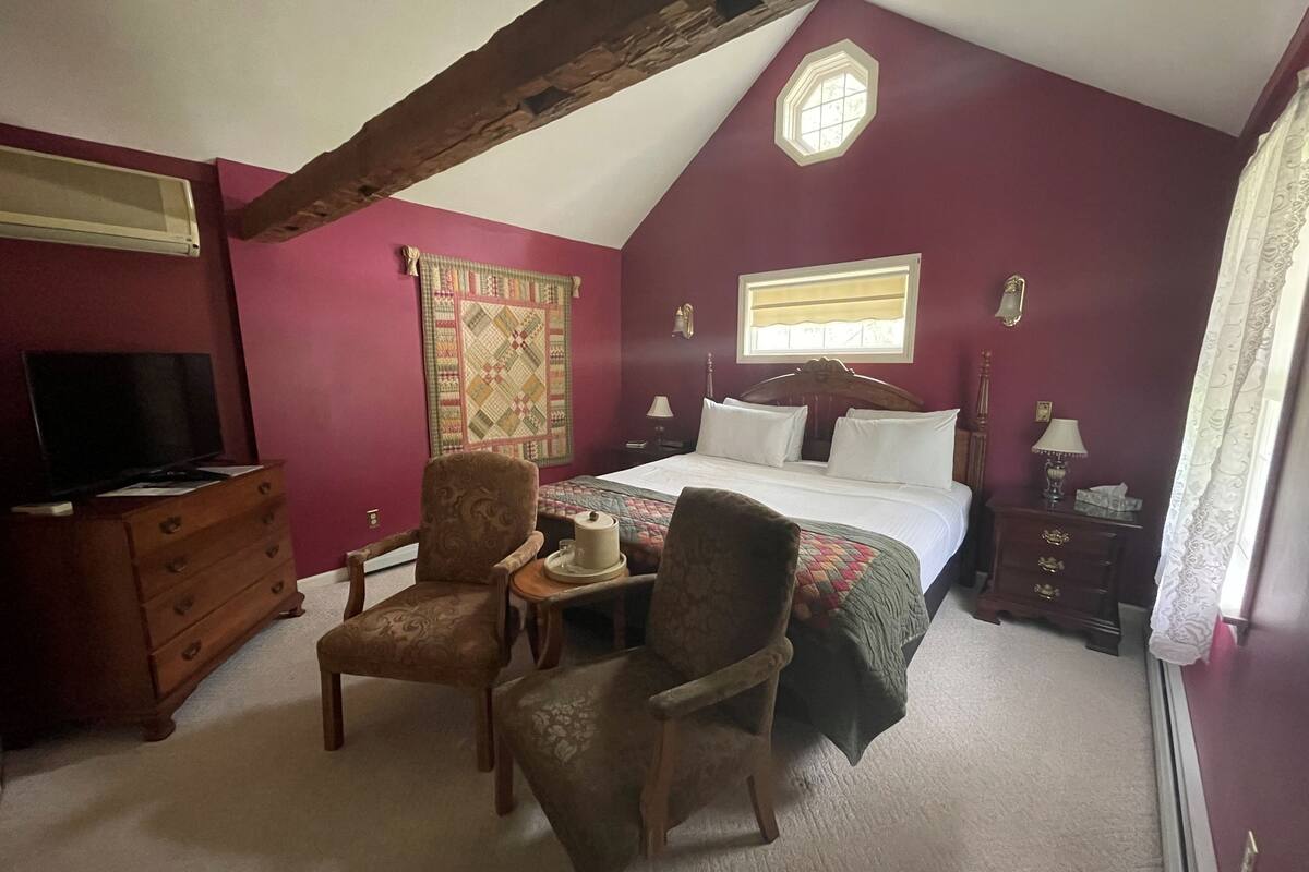 Une chambre à coucher comprenant un lit, deux chaises, une commode, un téléviseur et une fenêtre avec des rideaux.