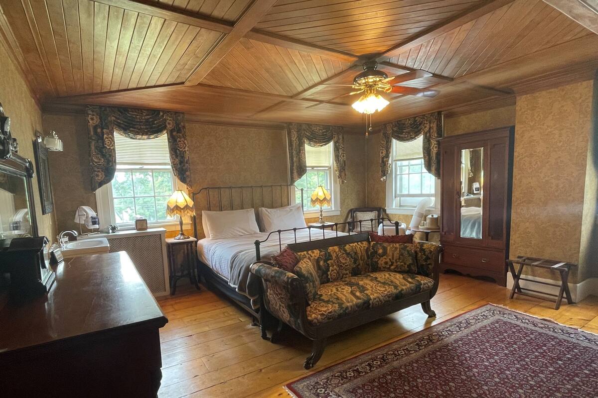 Une chambre avec un plafond en bois, un lit, un canapé vintage, une commode en bois et un grand tapis.