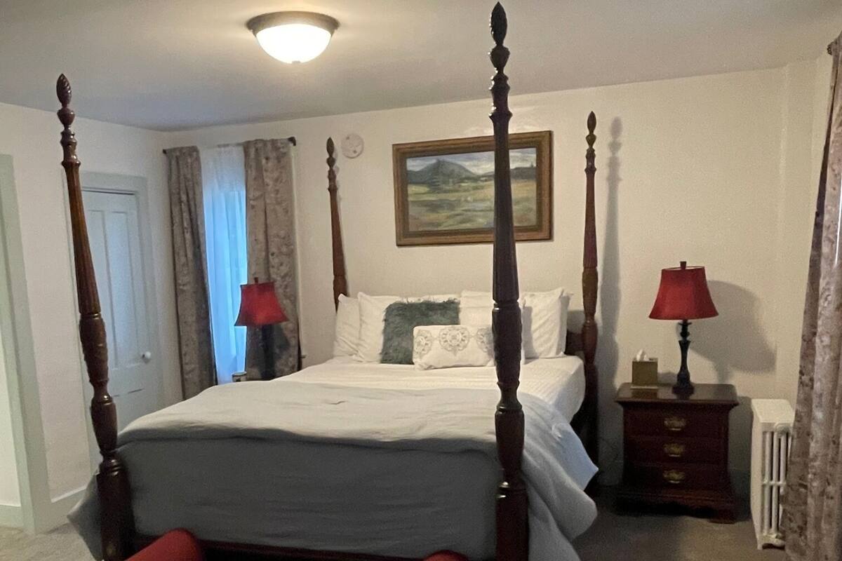 Un lit à baldaquin avec un banc placé devant, une table de chevet avec une lampe et un tableau accroché au mur.