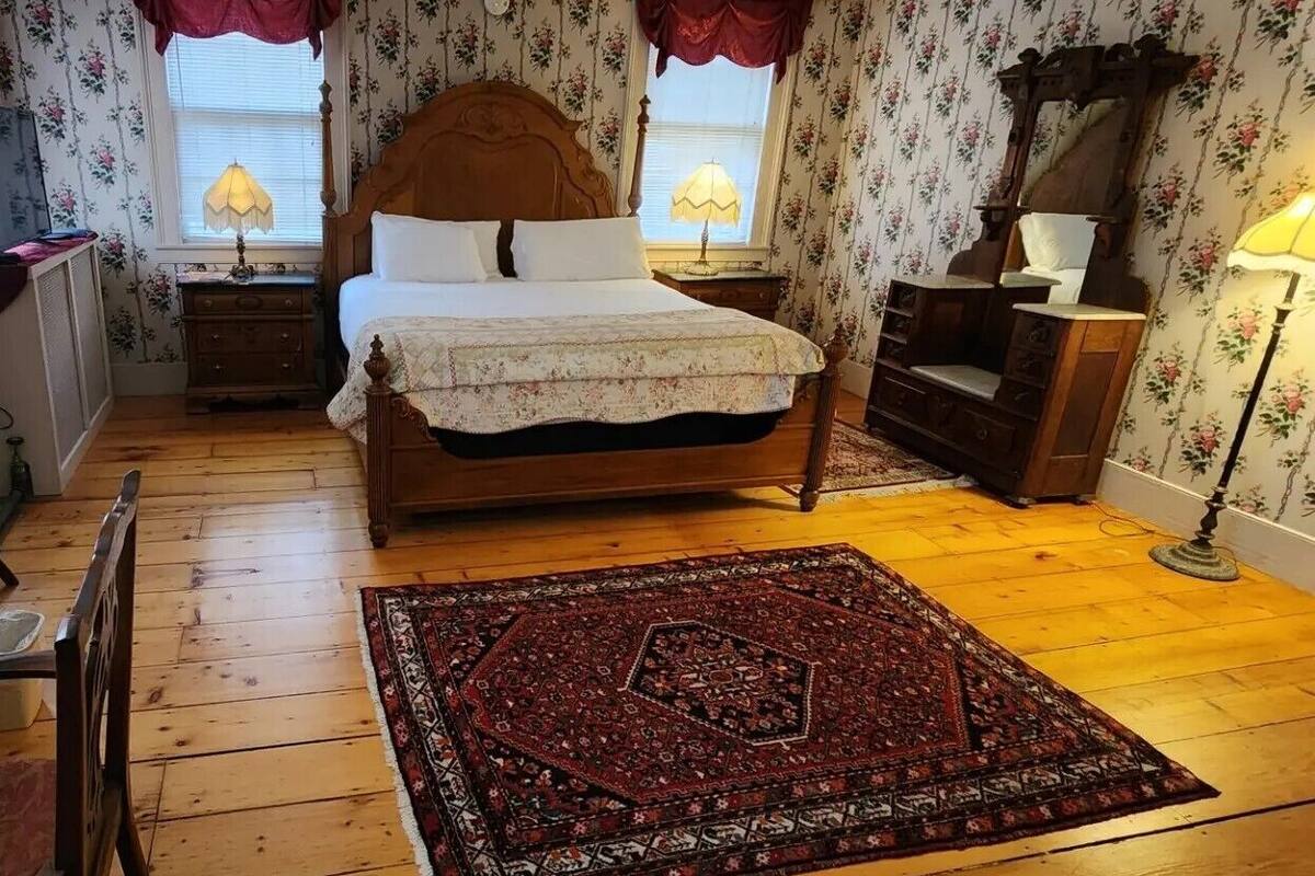 Une chambre avec un plancher en bois, un grand lit avec une tête de lit, une table de chevet, une commode et un tapis.