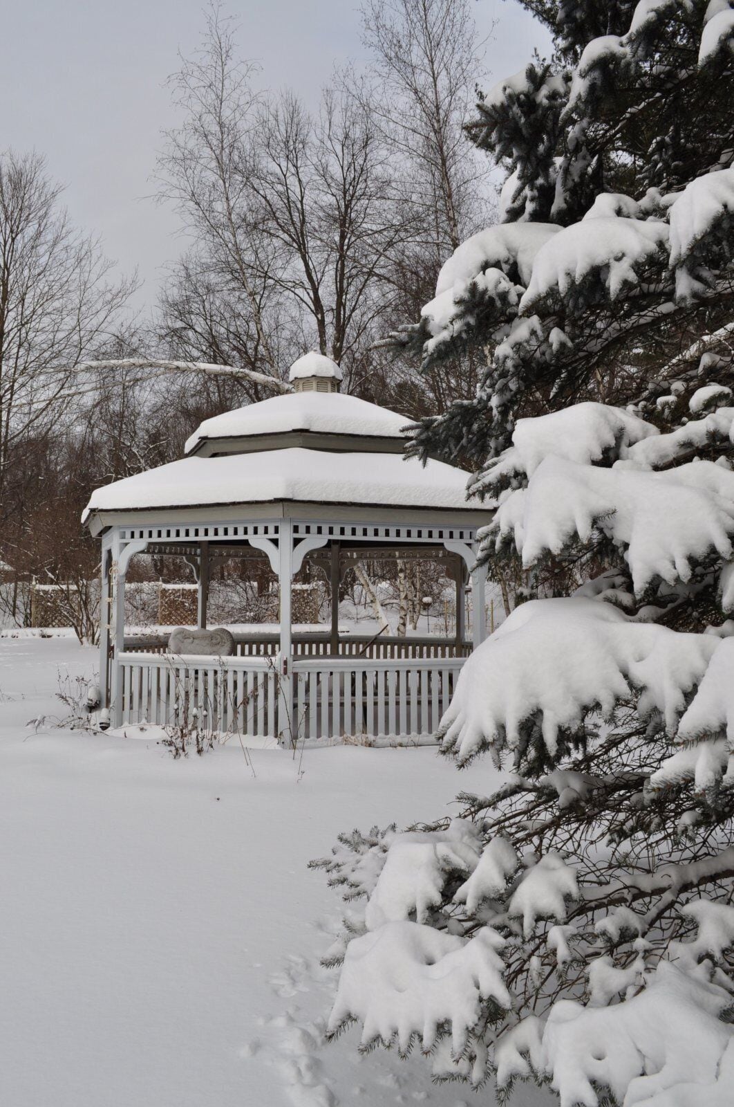 Ein mit Schnee bedeckter Pavillon, umgeben von schneebedeckten Bäumen und einem Zaun.