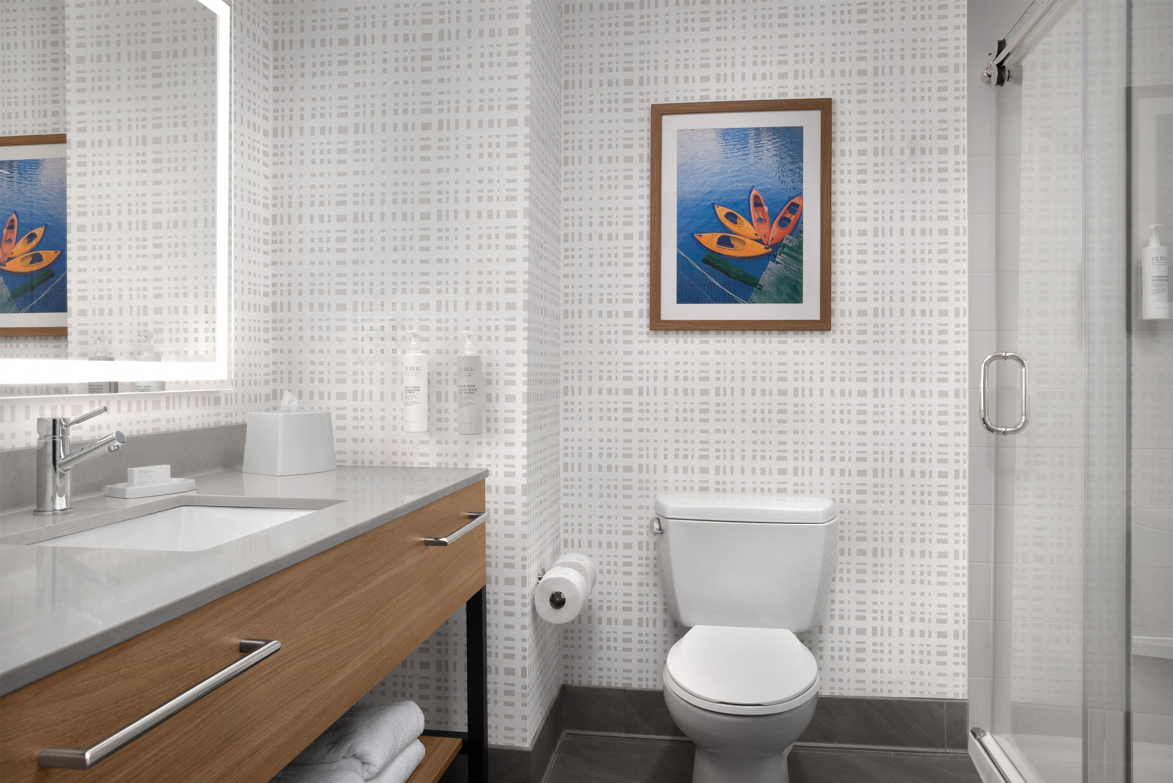 A modern bathroom with a white tiled wall, a wooden vanity, a white toilet, and a framed painting of kayaks.