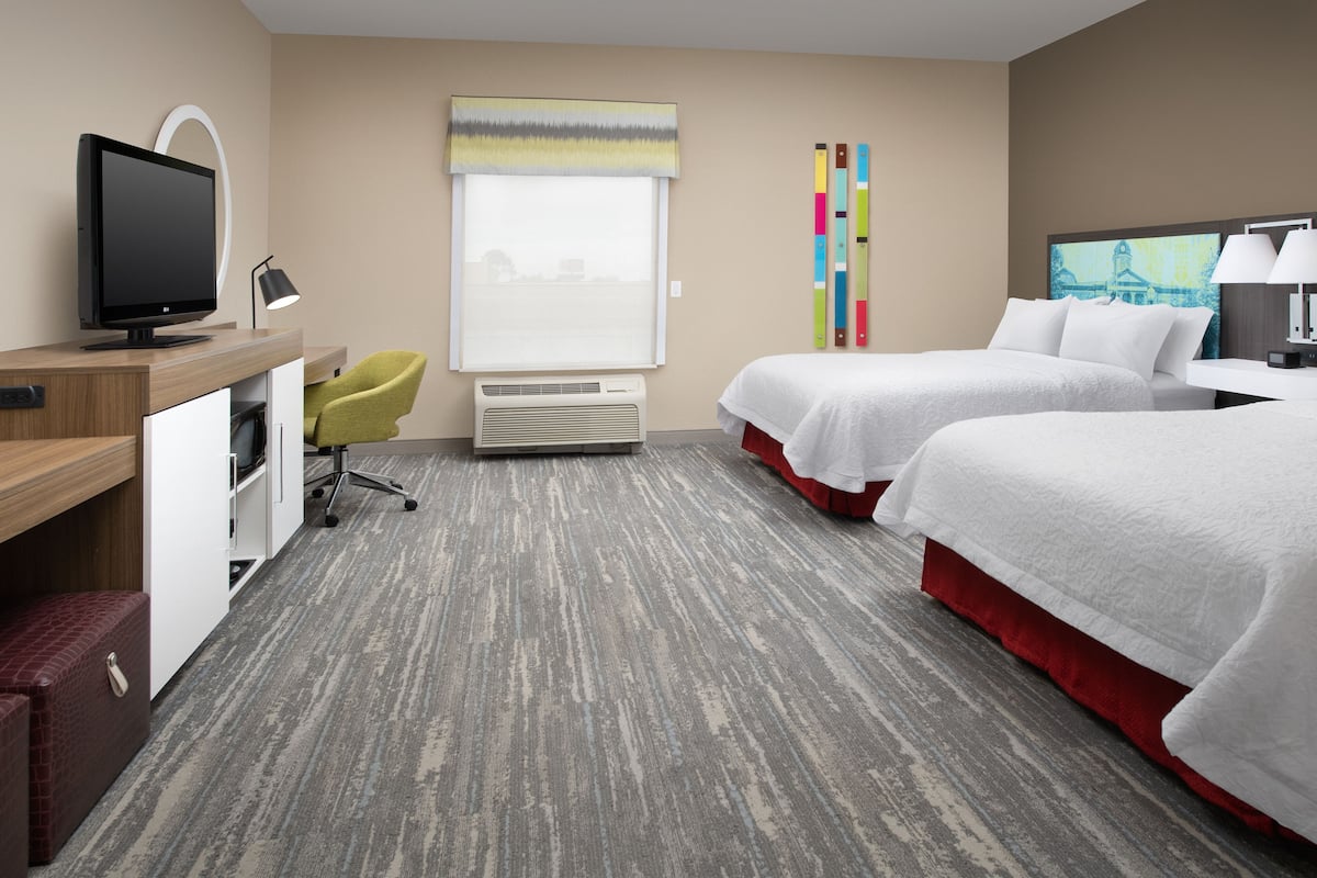 A hotel room with two beds, a desk with a computer, a chair, a window with blinds, and a wall-mounted lamp.