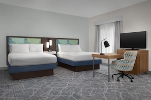 Habitación de hotel con dos camas, un escritorio, una silla, un televisor y obras de arte en las paredes.