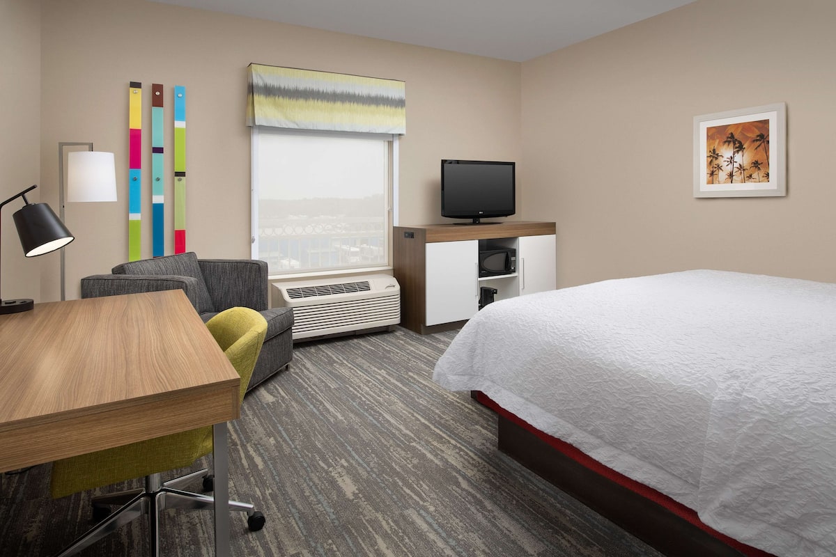 A hotel room with a bed, a desk, a chair, a television, a window with blinds, and a wall with a painting.