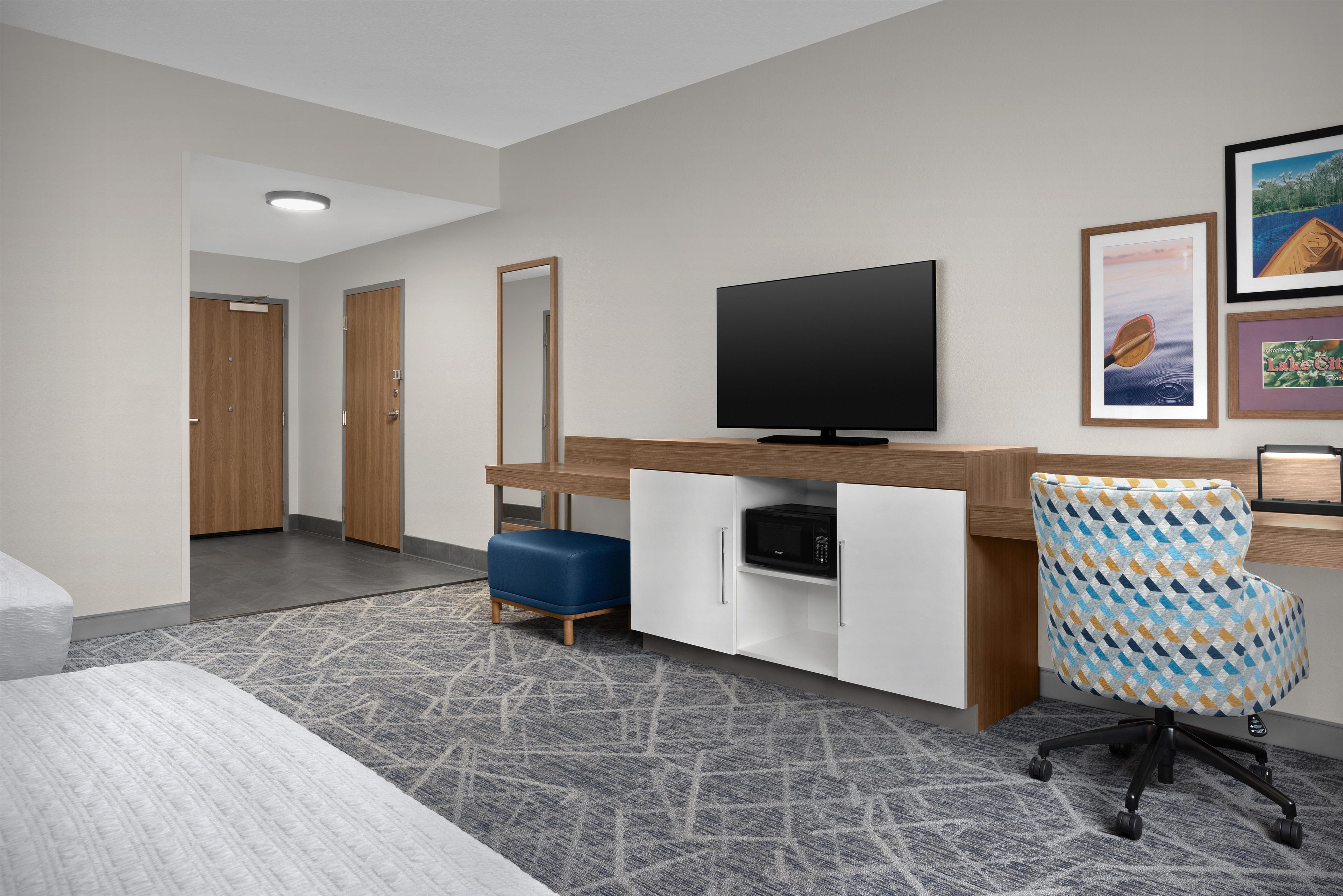 A hotel room with a flat-screen TV, a desk with a microwave, a patterned armchair, and a large mirror.