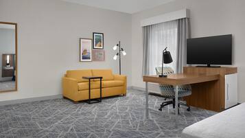 A hotel room with a sofa, a desk with a computer, a TV, a mirror, and framed pictures on the wall.