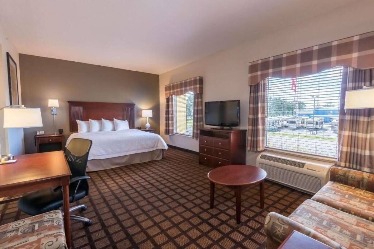 A hotel room with a bed, desk, chair, sofa, television, and large windows.