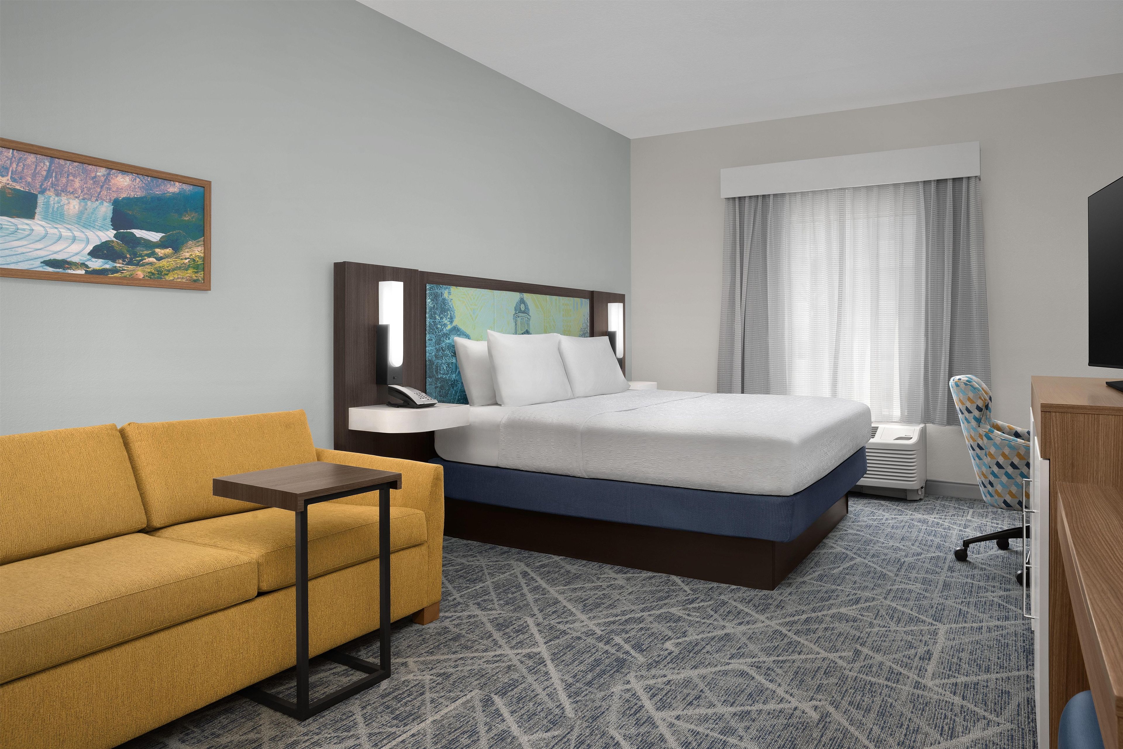 A hotel room with a bed, a sofa, a small table, a desk, a chair, and a painting on the wall.