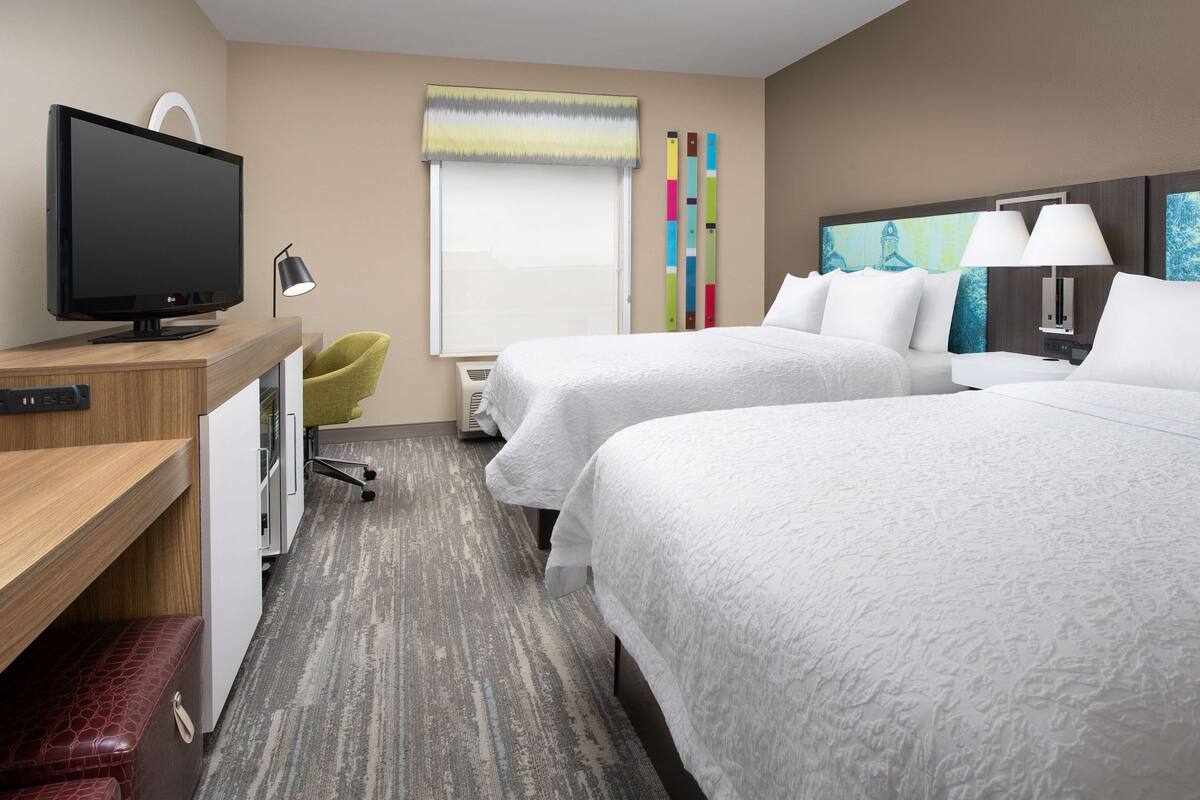 A hotel room with two beds, a desk with a television, a chair, and a window with blinds.