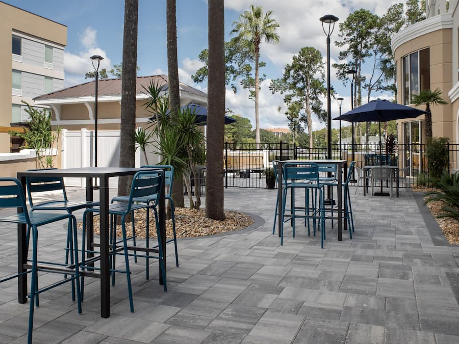 An outdoor patio area with tables and chairs, surrounded by palm trees and a building with large windows.