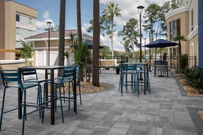 An outdoor patio area with tables and chairs, surrounded by palm trees and a building with large windows. - Hampton Inn & Suites Lake City (Lake City)