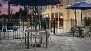 Eine Terrasse im Freien mit Tischen und Stühlen, umgeben von einem Metallzaun, mit Sonnenschirmen für Schatten.