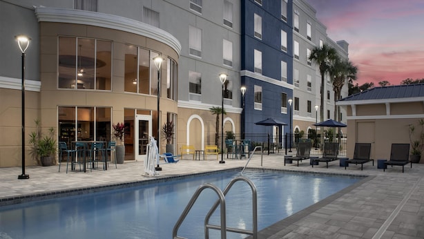 Ein Hotelpoolbereich mit Liegestühlen und Sonnenschirmen, ein Pool mit Metallgeländern sowie ein Gebäude mit mehreren Etagen und großen Fenstern.