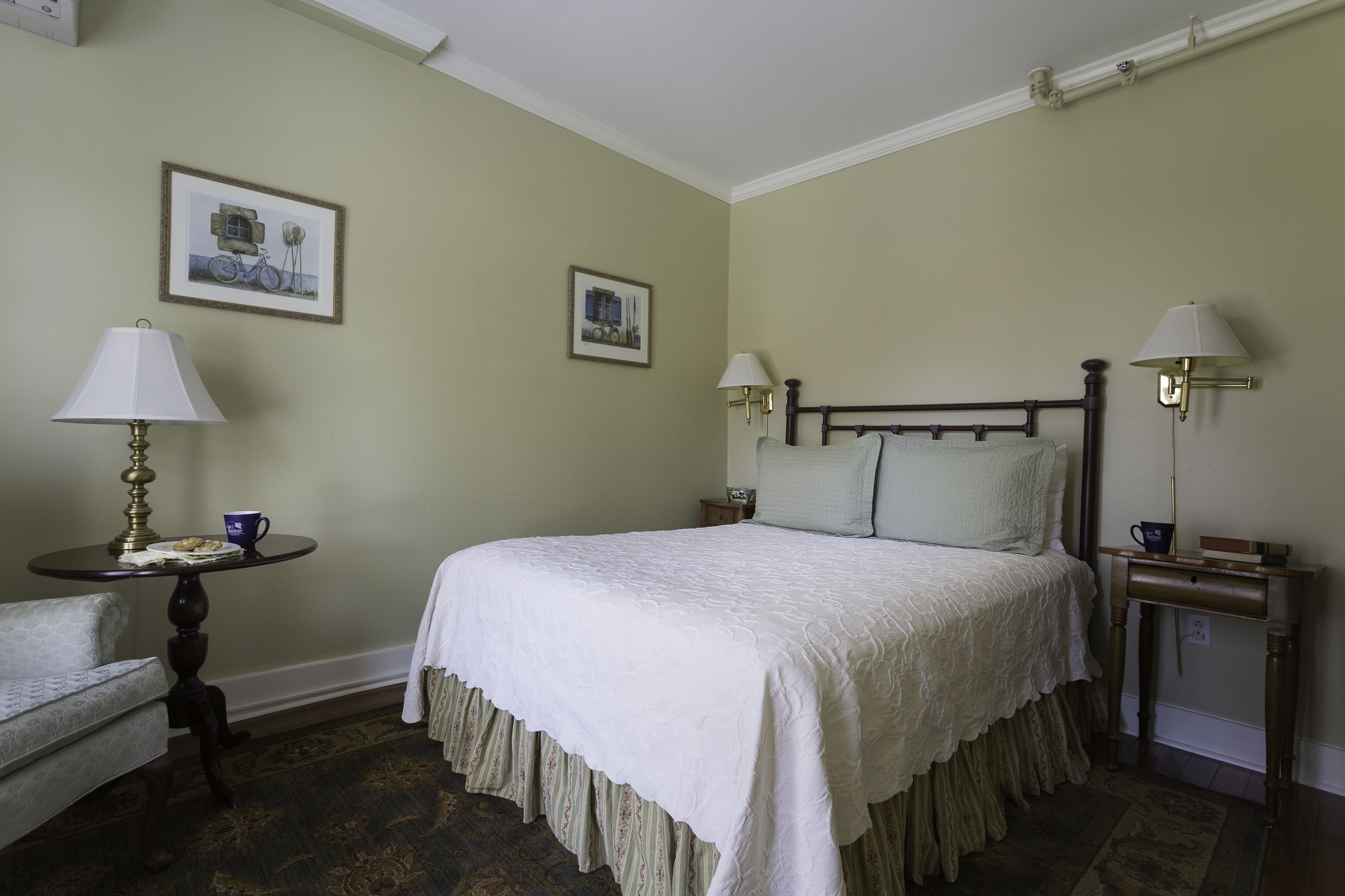 Un dormitorio ordenado con una cama grande, dos mesitas de noche con lámparas, una silla y dos cuadros en la pared.