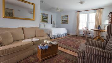 Una sala de estar con un sofá, un sillón, un cofre de madera y una mesita con una lámpara. Se observa una cama con colcha blanca y una ventana con cortinas.