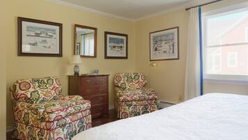 A bedroom with a bed, two patterned armchairs, a dresser, and three framed pictures on the wall.