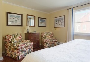 Un dormitorio con una cama, dos sillones estampados, un aparador y tres cuadros en la pared.