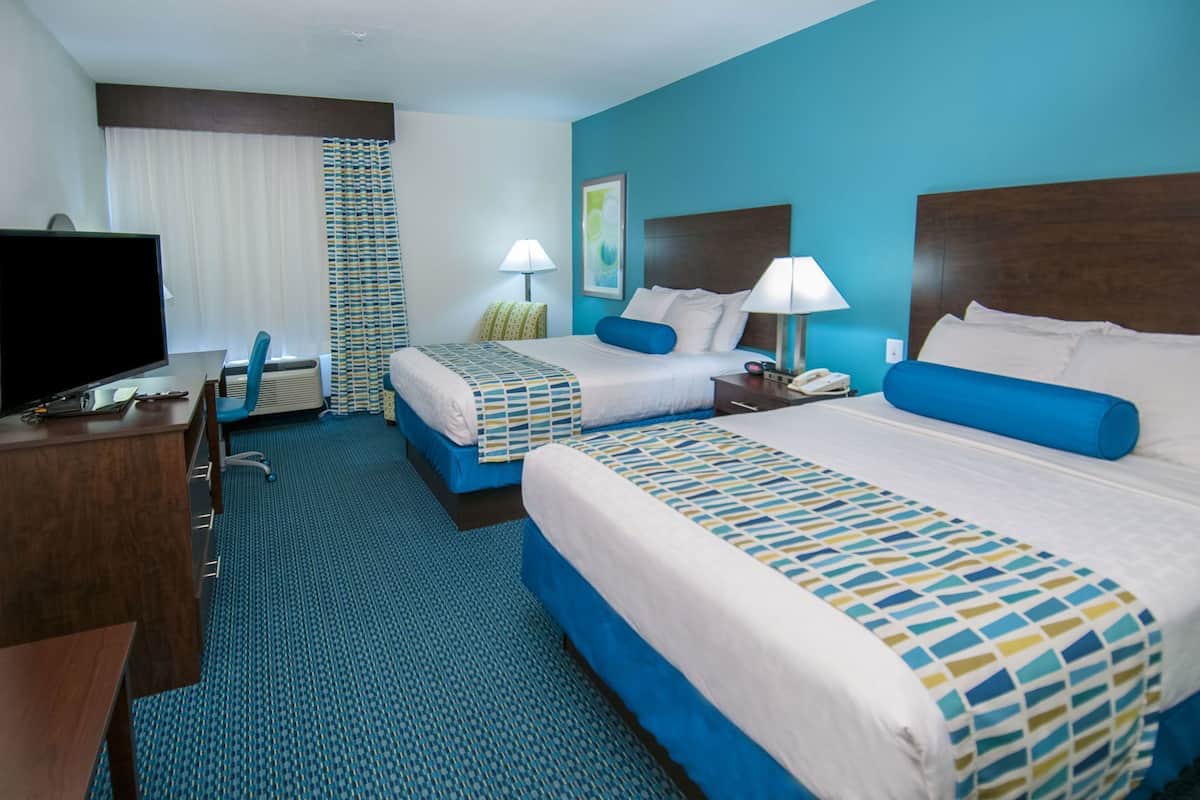 A hotel room with two beds, a desk, a chair, a TV, and a lamp.