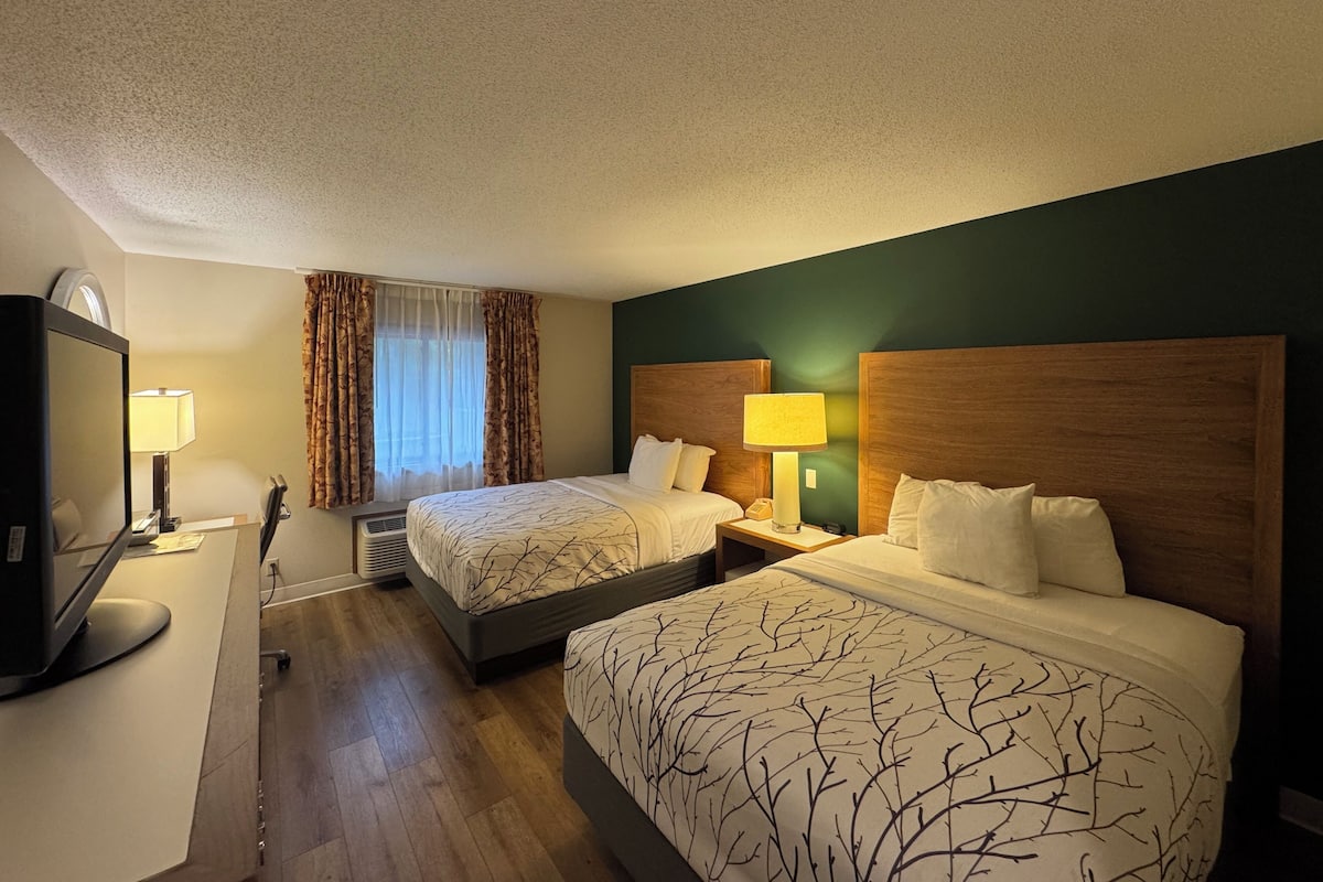 Quarto de hotel com duas camas, uma mesa de trabalho com computador, televisão e janela com cortinas.