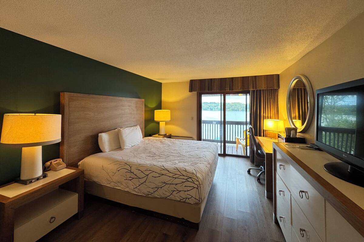 Quarto de hotel com cama, abajures, uma escrivaninha com televisão e vista para um lago pela janela.