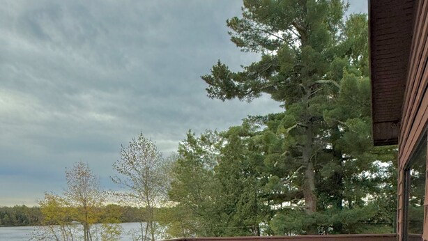 A covered deck with wicker furniture, a wooden table, and a view of a lake and trees. - St Croix Inn Solon Springs  (Solon Springs)