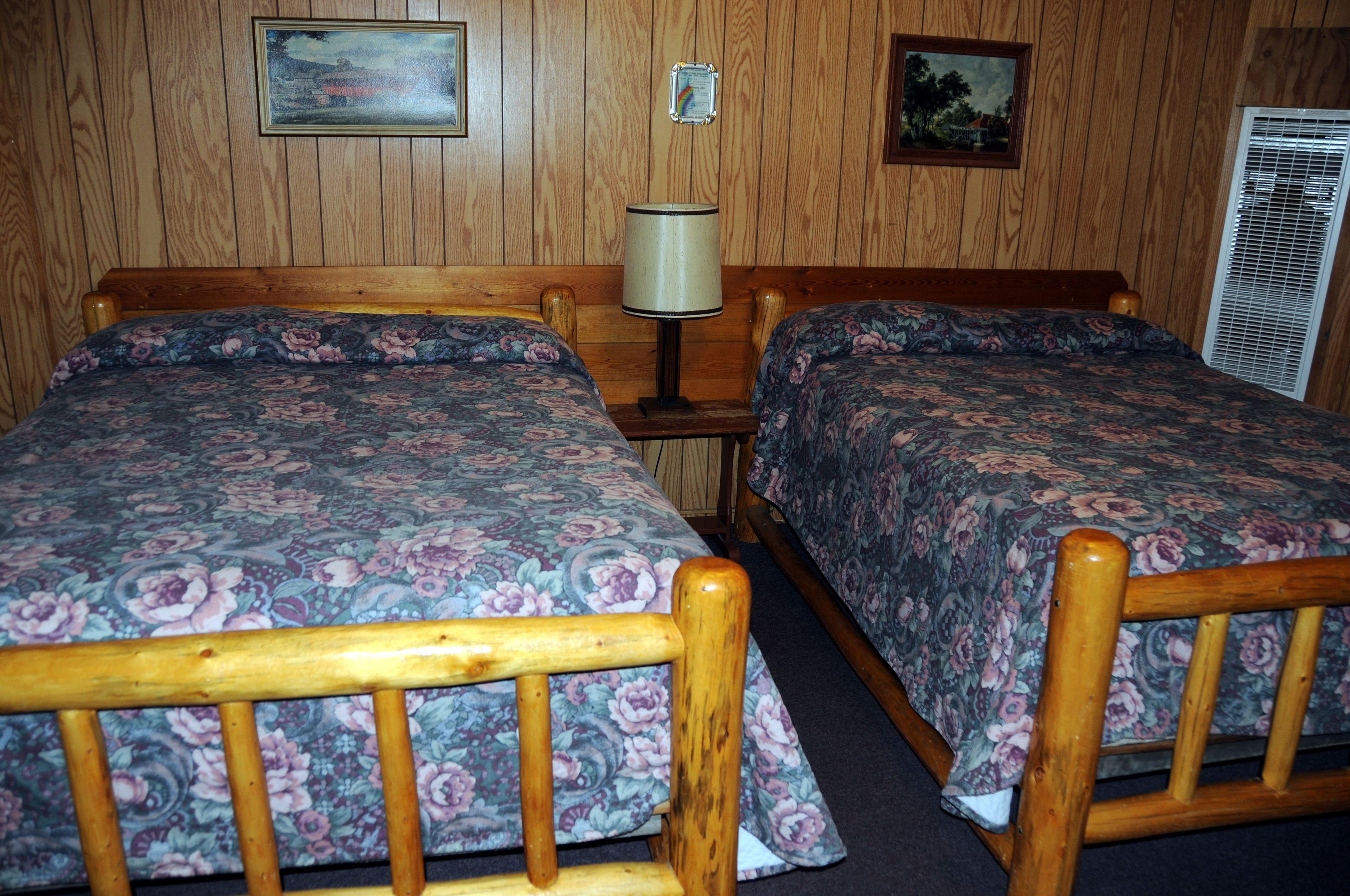 A room with two wooden beds with floral bedding, a wooden nightstand with a lamp, and a wall-mounted air conditioning unit.