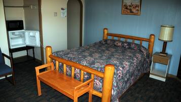 A small, compact room with a single bed, a wooden bench, a small table, a refrigerator, and a nightstand with a lamp.