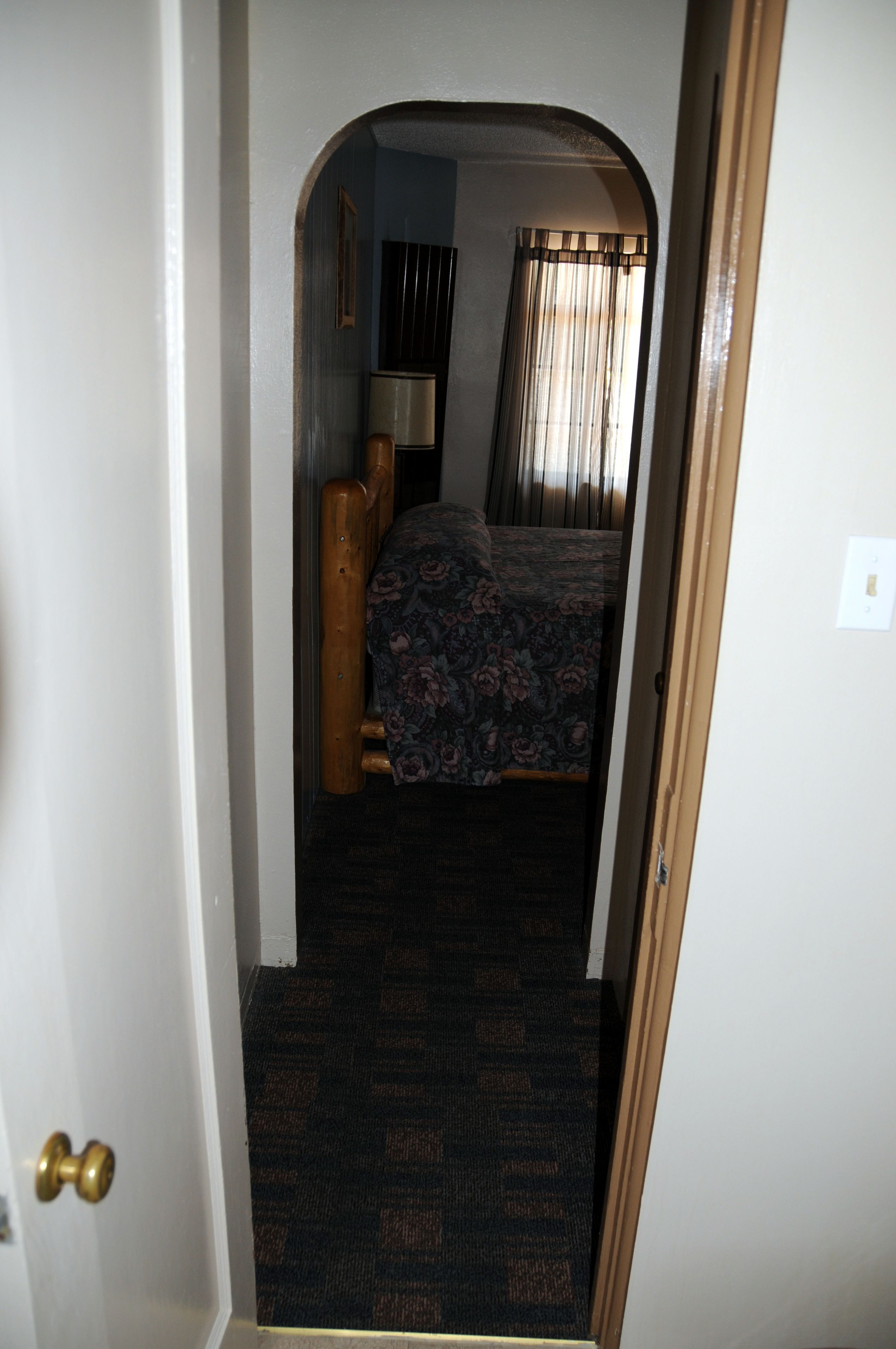 A narrow hallway leading to a bedroom with a bed, a wooden headboard, and a window with curtains.